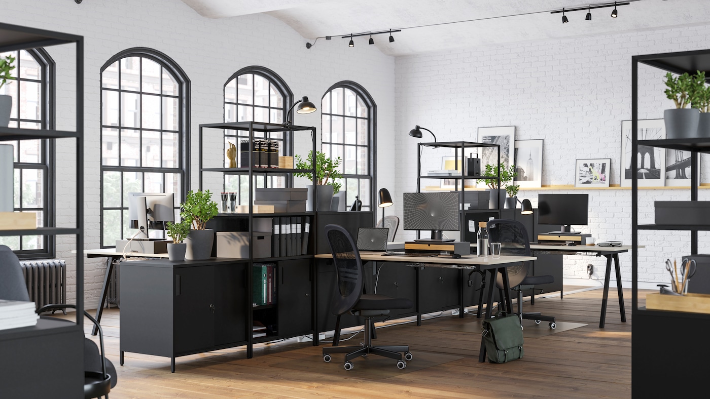 A bright open-plan office space divided using black TROTTEN cabinets and open shelving filled with plants, files and boxes.