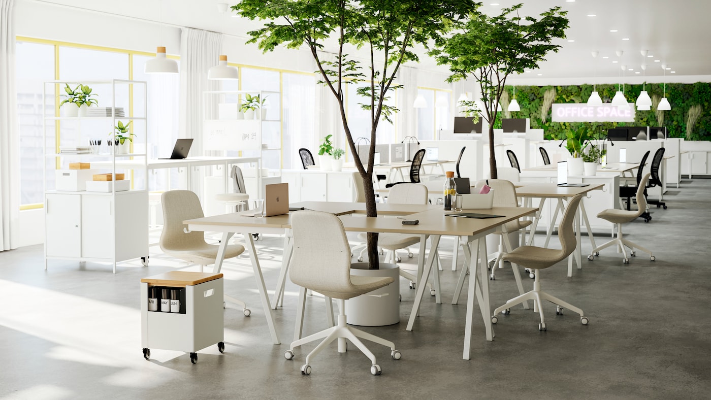 An open office with several groups of beige/white desks and chairs put together as meeting spots, green plants in between.