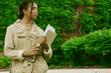 a woman in a tan jacket holds a notebook and a piece of paper in front of a cbs logo