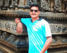 a man wearing sunglasses and a blue shirt is standing in front of a stone wall