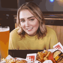 a woman in a yellow sweater sits at a table with a plate of food and a glass of beer with the word delish on it