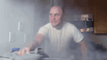 a bald man is ironing clothes in a kitchen while smoke is coming out of the stove .