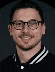 a man wearing glasses and a black and white varsity jacket smiles for the camera