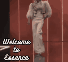 a woman in a fur coat is standing in front of a red wall with the words welcome to essence below her