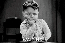 a black and white photo of a little boy sitting at a desk with his hand on his chin and the words `` waiting '' .