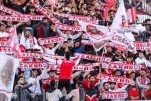 a man in a red shirt stands in a crowd holding up a zakho scarf