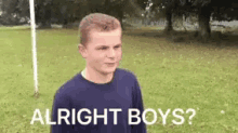 a young man is standing in a grassy field with the words `` alright boys '' written on the ground .