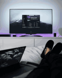 a person is laying on a bed with their feet up in front of a flat screen tv that is playing hustle by another