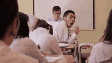 a group of students are sitting at desks in a classroom while a man stands in front of them .