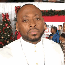 a man with a beard and a white shirt is standing in front of a christmas tree and looking at the camera .