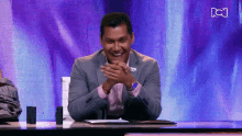 a man in a suit sits at a desk in front of a purple screen with the letters bc on it