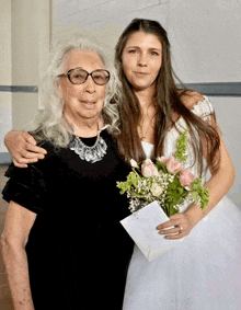 a woman in glasses stands next to a woman in a white dress holding flowers