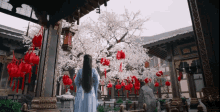 a woman in a blue dress is standing in front of a tree with red lanterns hanging from it .