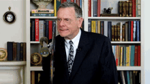 a man in a suit and tie stands in front of a bookshelf with a book titled the weatherman on top