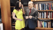 a man in a suit and tie is talking to a woman in a yellow dress in front of a bookshelf