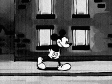 a black and white cartoon of mickey mouse walking down a street in front of a building .