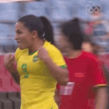 a woman in a yellow soccer jersey is running with her fist in the air while another woman in a red shirt watches .