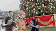 a man and a woman wearing masks are standing next to a gingerbread man in front of a christmas tree .