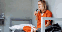 a woman in an orange prison uniform is sitting on a bed eating a candy bar .