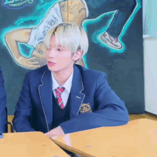 a young man in a school uniform and tie is sitting at a table in front of a painting .