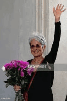 an older woman is holding a bouquet of purple flowers and waving .