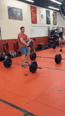 a man in a red shirt is lifting a barbell in a gym with a us navy seals poster on the wall