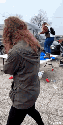 a woman in a gray jacket is standing next to a woman in a white shirt who is jumping on a table