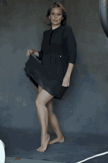 a woman in a black dress stands barefoot in front of a grey background