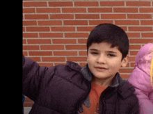 a young boy wearing a purple jacket is standing in front of a brick wall