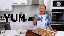 a woman in a blue tie dye shirt is eating a cookie in a kitchen with the word yum behind her