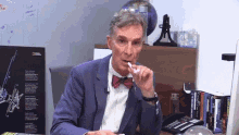 a man in a suit and bow tie is sitting at a desk with a national geographic poster in the background