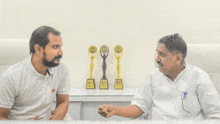 two men are sitting in front of a table with trophies on it one of which says ' winner ' on it