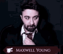 a man in a suit and tie is sitting in front of a microphone with the name maxwell young on it .