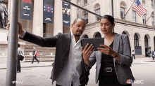 a man and a woman are looking at a tablet in front of a building with the nbc logo on it