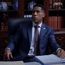 a man in a suit and tie is sitting at a desk in front of a bookcase that says bet on it