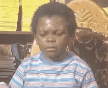 a young boy in a striped shirt is sitting in front of a bookshelf and making a funny face .