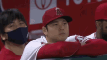 a baseball player wearing a red hat and a white shirt is sitting in the dugout with his arms crossed .