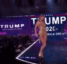 a woman in a gold dress is singing into a microphone on a stage with a trump sign in the background .