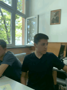 a boy in a black shirt sits at a desk in front of a window with a picture of a man on the wall above him