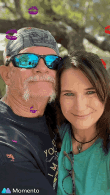 a man wearing sunglasses and a bandana stands next to a woman wearing a momento t-shirt