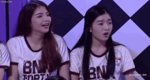 two girls are sitting next to each other in front of a checkered wall wearing bnk shirts .