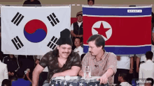 two men sit at a table in front of a korean and north korean flag ..