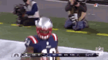 a football player in a patriots uniform is kneeling on the field while a photographer takes a picture of him .