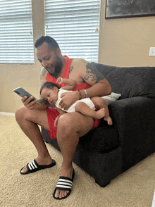 a man is sitting on a couch holding a baby and looking at a cell phone