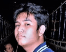 a young man in a blue jacket is standing in front of a fence and looking at the camera .