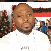 a man with a beard and a necklace is standing in front of a christmas tree and talking to the camera .