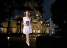 a woman in a pink dress is holding a book in front of a building at night