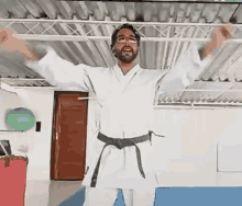 a man in a white karate uniform with a black belt is standing with his arms outstretched in a gym .