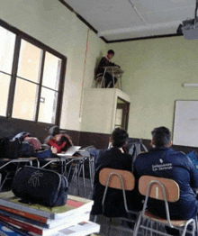 a man sits at a desk in a classroom wearing a jacket that says jalisco on the back