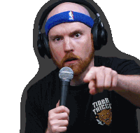 a man wearing headphones and a blue headband holds a microphone and points at the camera